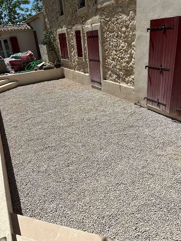 Espace vert terminé avec gravier sur un terrain préparé, créant une surface prête pour l'aménagement extérieur, autour d'une maison en pierre avec fenêtres à volets rouges.