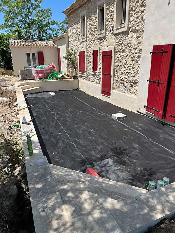 Terrain préparé avec géotextile pour l'aménagement d'un espace vert, avant la pose de gravier, près d'une maison en pierre avec fenêtres à volets rouges.